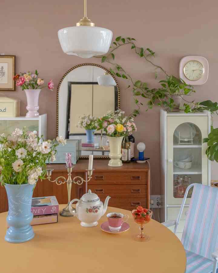 A living room with soft pink walls, featuring a yellow dining table with vases, candelabra, and a wall clock in vintage style