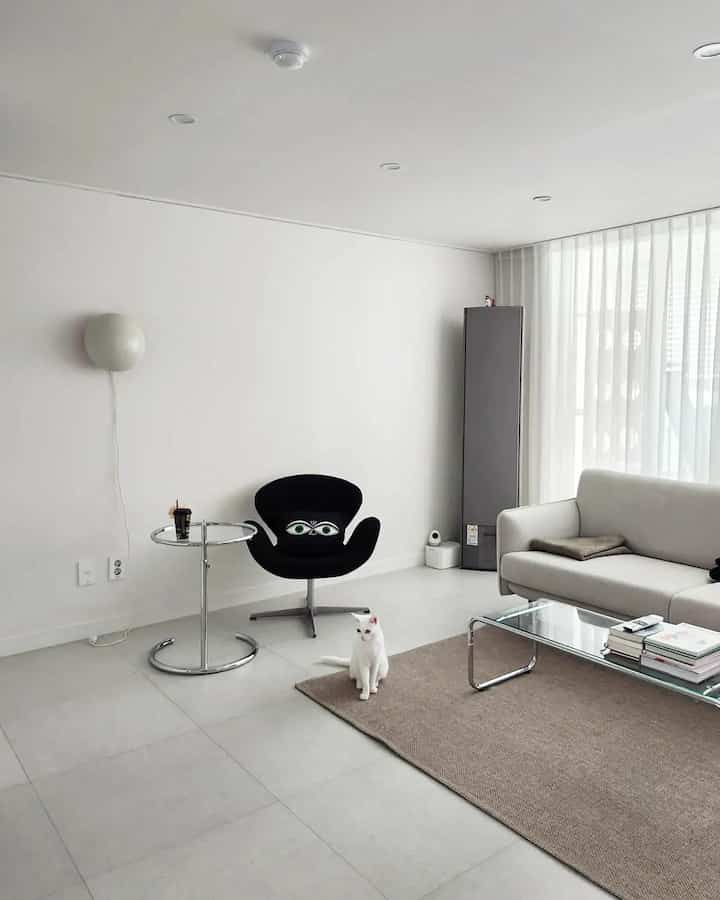 Minimal white-toned living room featuring light beige sofa, black armchair, glass coffee table, and a white cat in a clean interior