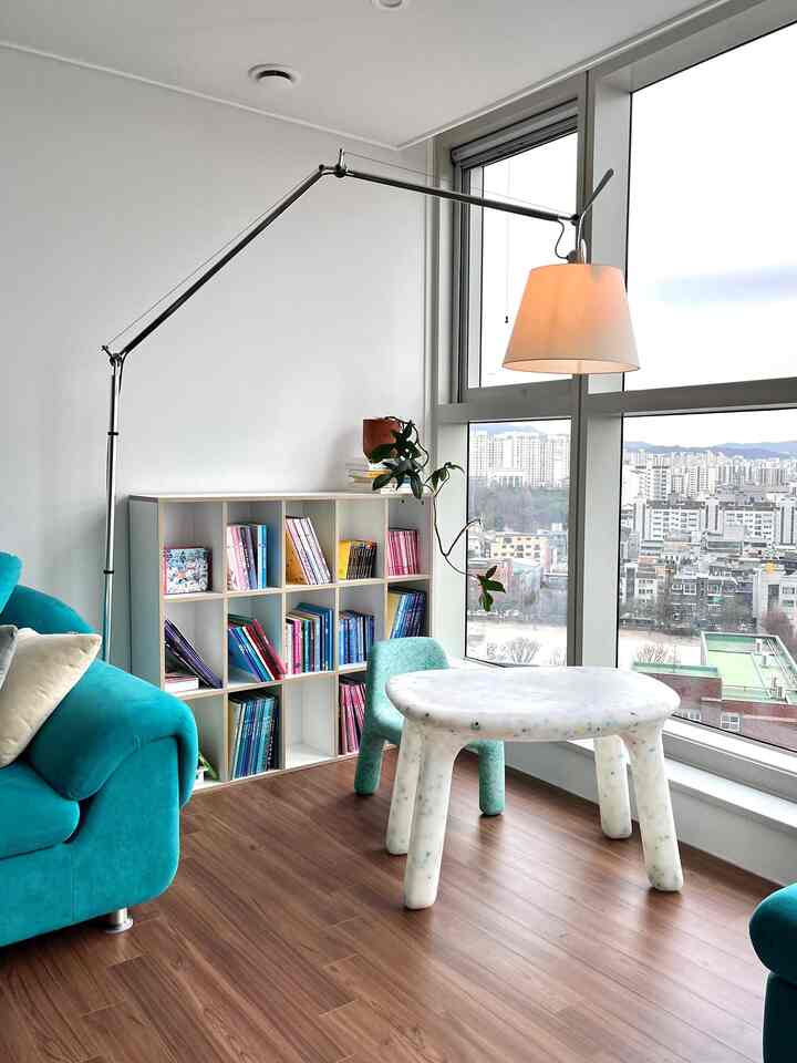 Bright living room corner in teal tones featuring a baby study with floor-to-ceiling windows, bookshelf, wooden floor, and floor lamp creating a natural atmosphere