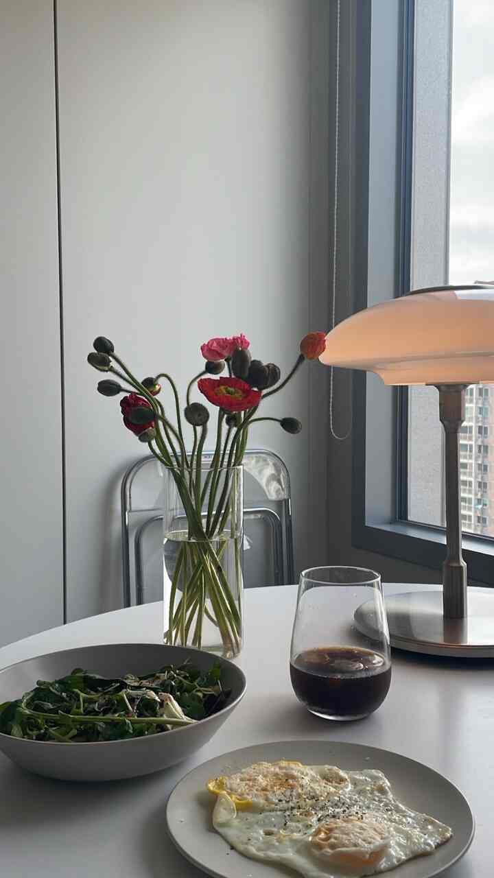 Minimal dining space in bright white and beige tones, featuring transparent folding chair, vase with flowers, and a neat breakfast setup