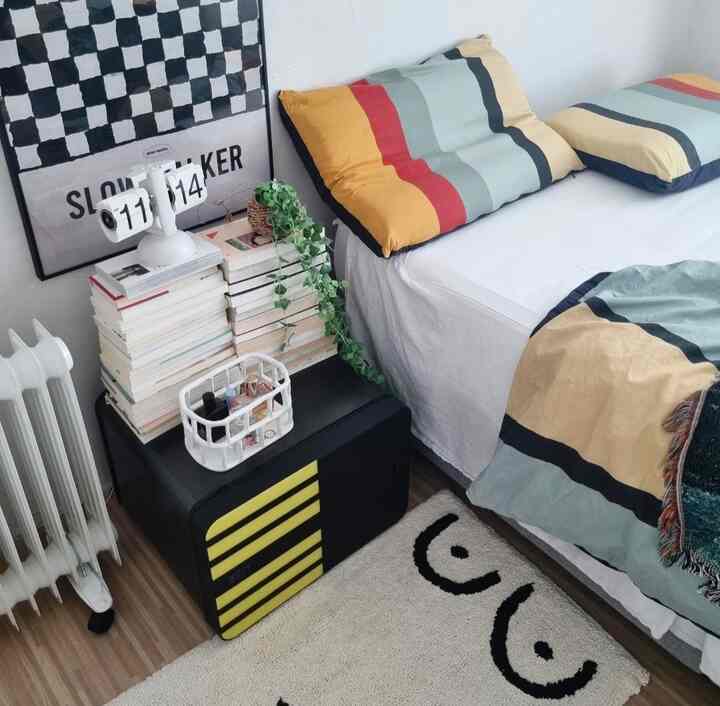 White and vintage-toned bedroom featuring a cozy bed with colorful pillows, a nightstand stacked with books and decorative items