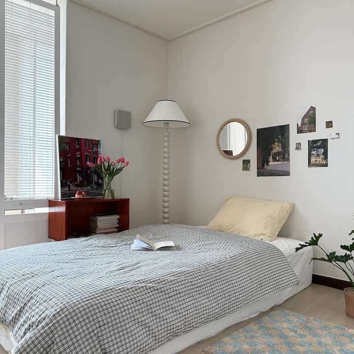 A white and gray toned bedroom featuring a bed with checked duvet cover, floor lamp, and wooden framed round mirror in a simple natural setting