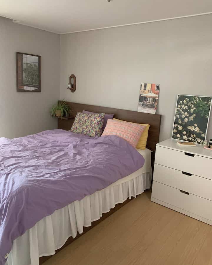 ナチュラルな色調の寝室、柔らかい紫のベッドカバーと花柄クッション、白いドレッサーと木目調のヘッドボードが調和した雰囲気のあるインテリア