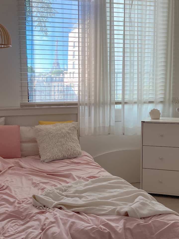 ホワイトとピンクの色調の子供部屋、ピンクの寝具と白いチェスト、木製ブラインドが配置された明るく整った雰囲気のあるインテリア