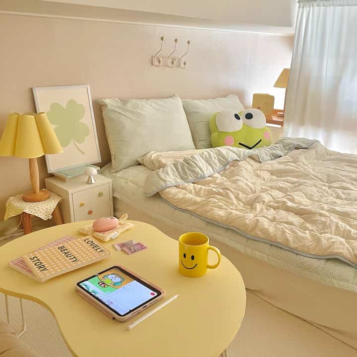 Cozy bedroom in soft beige and light green tones featuring a cute frog cushion and yellow table lamp creating warm ambiance
