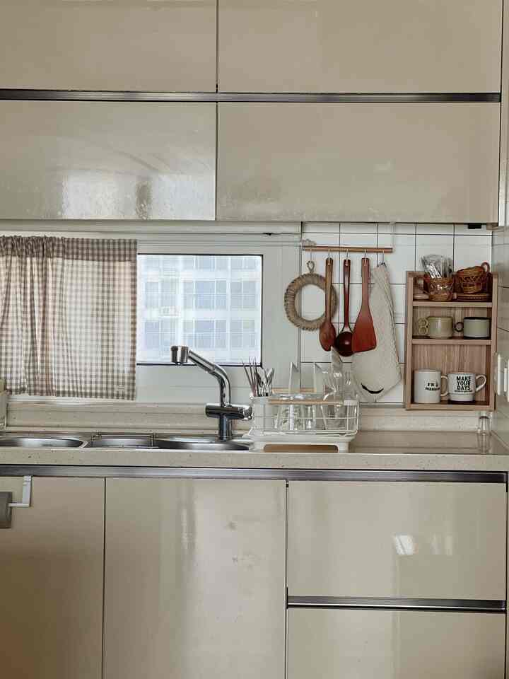 A compact kitchen space in beige and white tones featuring a gingham curtain on the window and wooden kitchen utensils, clean and minimalist interior