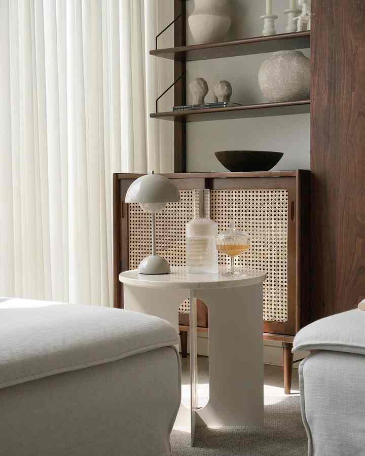 Natural-toned living room in white and wood tones, featuring a central side table with table lamp and rattan cabinet