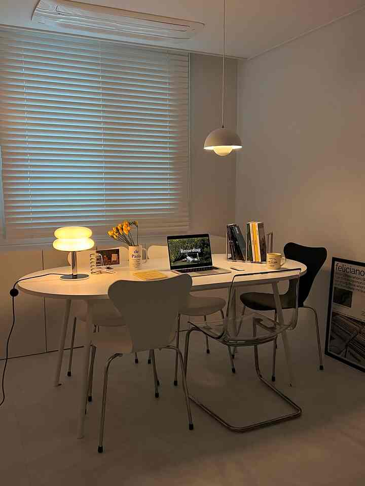 White-toned dining room featuring a long central table, assorted chairs, laptop, and lamps creating a clean modern atmosphere