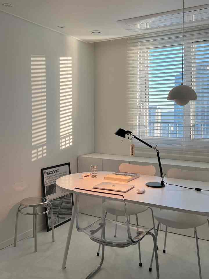 White-toned study featuring a central desk, transparent chair, stool, and table lamp in a modern setting
