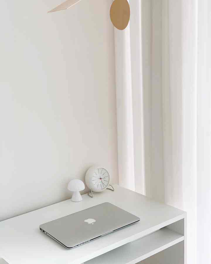 White-toned home office space featuring a white desk with a silver MacBook, small clock, and table lamp, creating a clean workspace