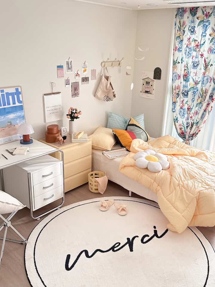 暖かいイエローとホワイトのトーンの一人暮らし寝室、ベッドとデスク、黄色のチェスト、花柄のカーテンが配置された雰囲気のある空間