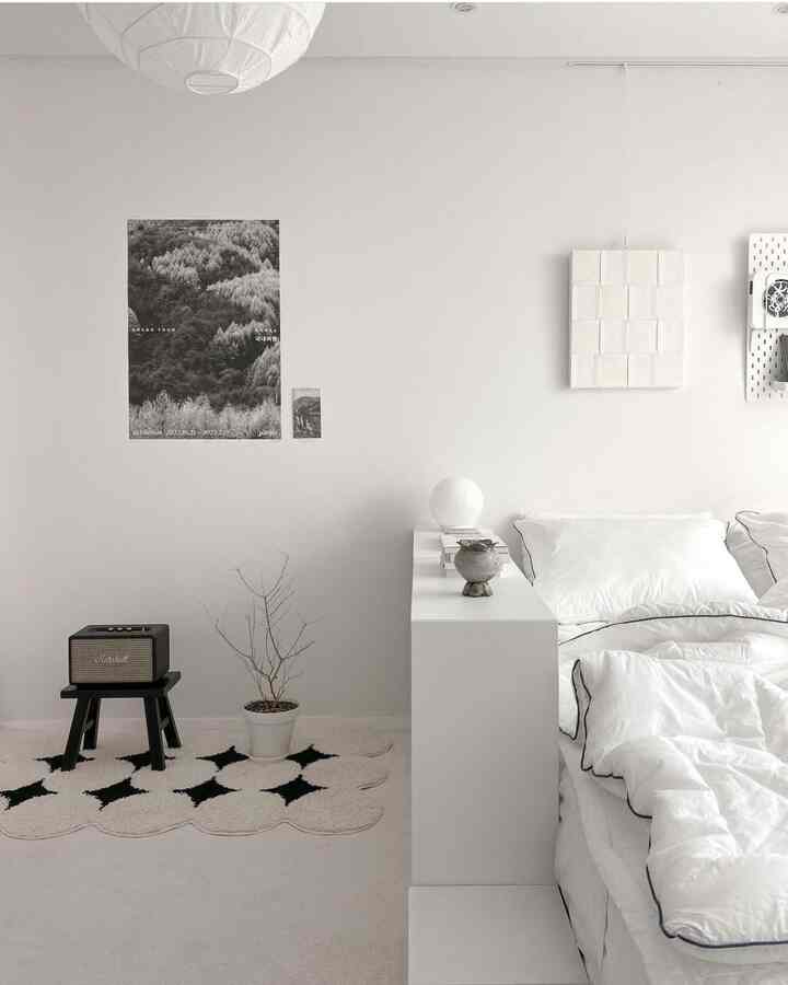 White-toned bedroom featuring a white bed and side table, black and white posters, and a patterned rug in a simple setting