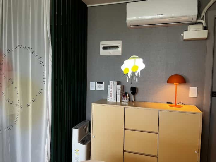 グレーの壁とイエローの収納棚が配された玄関隣接スペース、ディープグリーンの間仕切りと白いカーテンが調和した温かみのあるインテリア