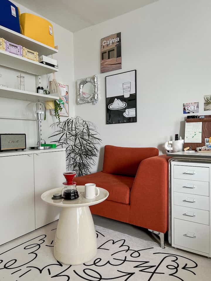 White and red-toned study space featuring a round table with drip coffee set at center and an orange sofa, creating a cozy home cafe atmosphere