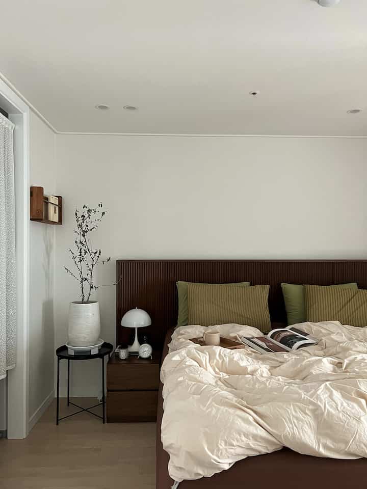 Beige and brown toned bedroom featuring a wooden bed, nightstand, white table lamp, and a potted plant creating a calm space