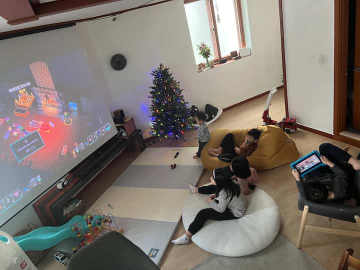 Small bedroom with white walls and wood-toned floor featuring kids, a bean bag, LED Christmas tree, and a large game screen creating a cozy play and home cinema space