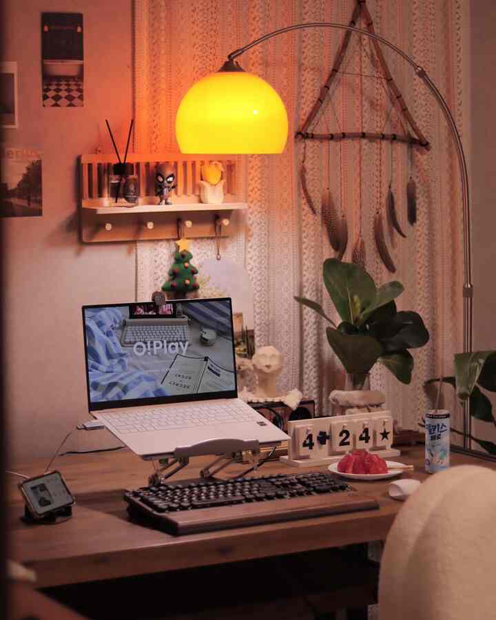 Warm wood-tone home office desk featuring laptop on stand, Bluetooth keyboard, decorative shelf, and glowing floor lamp creating cozy atmosphere
