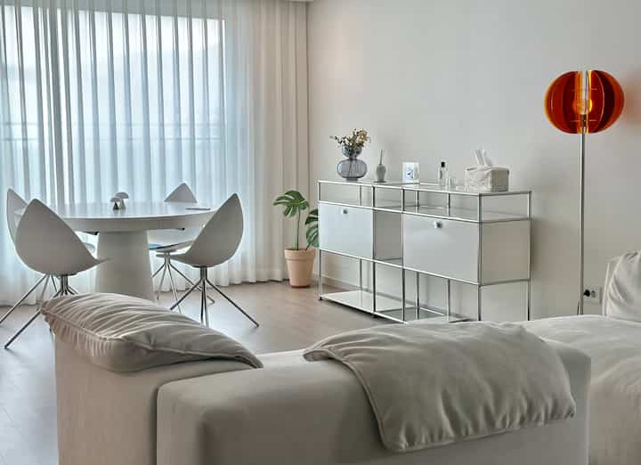White-toned living room featuring a modular sofa, round dining table, and storage cabinet in a clean modern setting