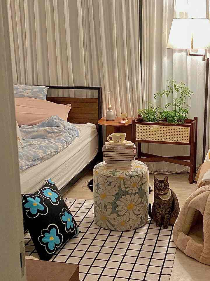 ベージュ色のカーテンと木製家具がある寝室、猫と花柄のスツールが配置された温かみのあるインテリア