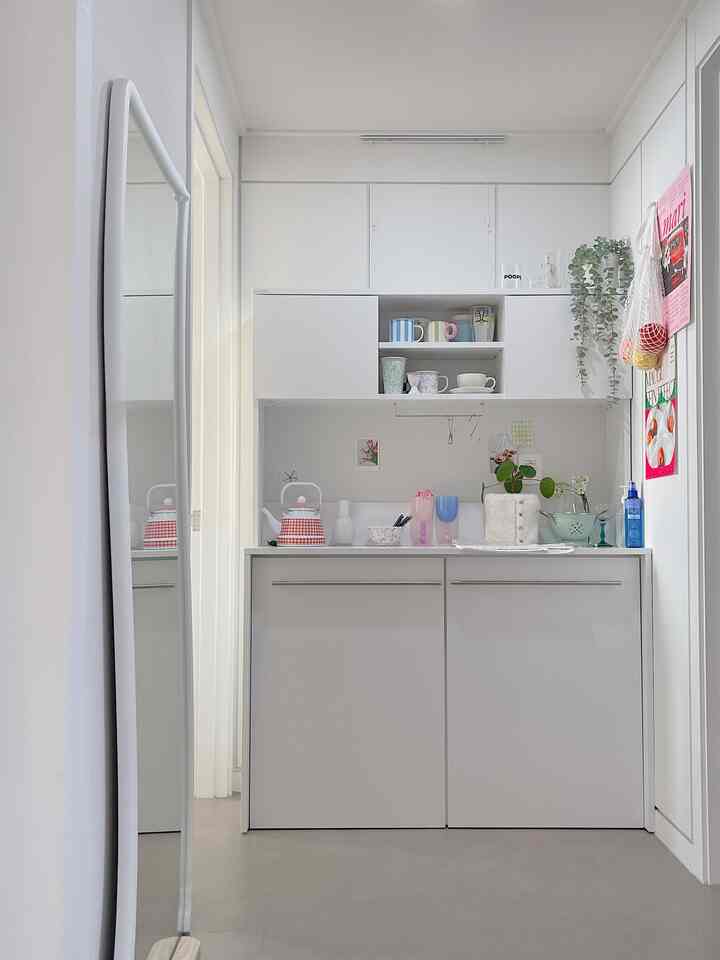 White-toned kitchen space featuring neat storage cabinet, various cups, and plants creating cozy home cafe atmosphere
