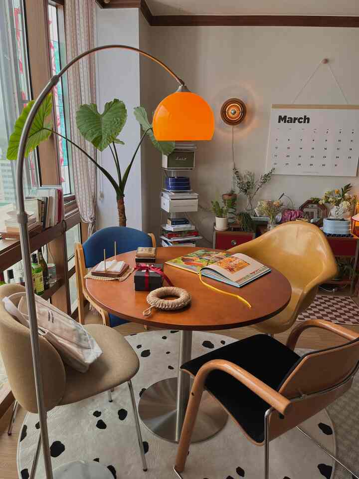 Living room with orange lighting, round dining table, various chairs, and large plants in a vintage mid-century modern style