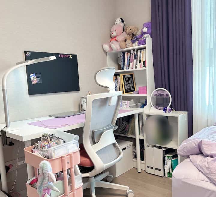 Lavender and white tone bedroom home office with tidy desk, ergonomic chair, bookshelf, and curtains creating a calm atmosphere