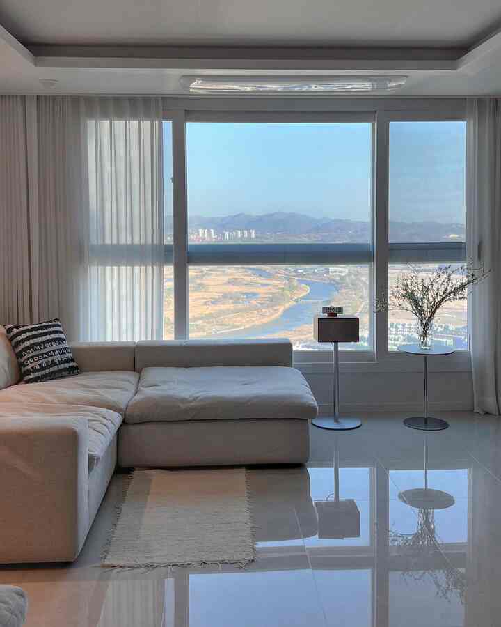 明るい白を基調としたリビング、コーナーソファとシフォンカーテンが配置され、大きな窓の向こうに川と山の絶景が広がる雰囲気のあるインテリア。