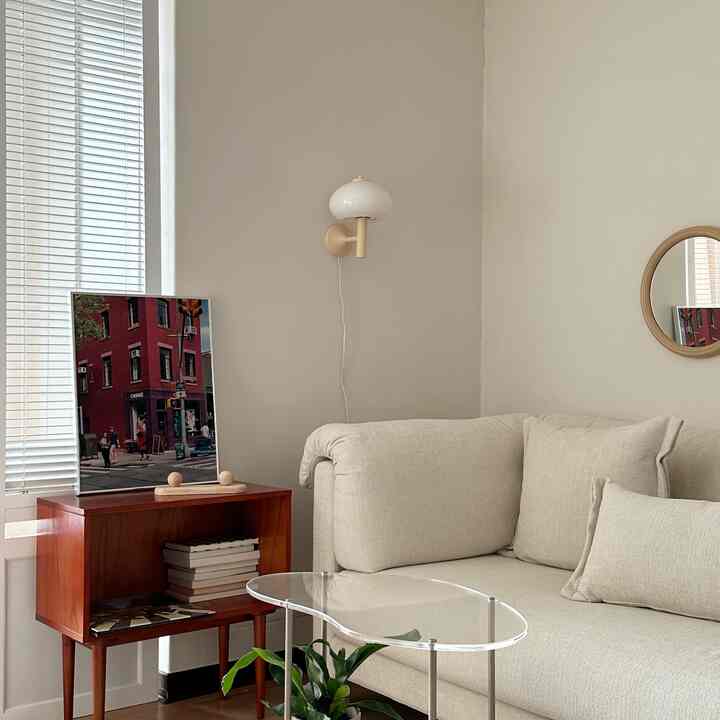 Soft beige-toned living room featuring a comfortable sofa, transparent coffee table, wooden side table, and wall sconce creating a cozy atmosphere
