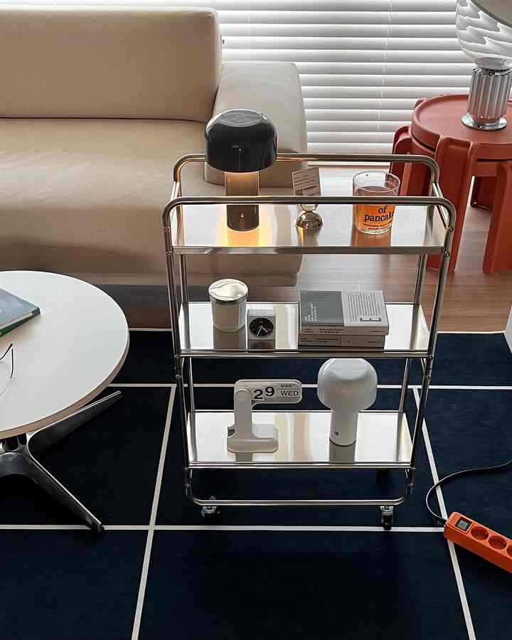 ベージュのソファとネイビーのラグの上にステンレス製の3段ワゴンが置かれた、清潔感のあるモダンなミッドセンチュリーモダンのリビング空間