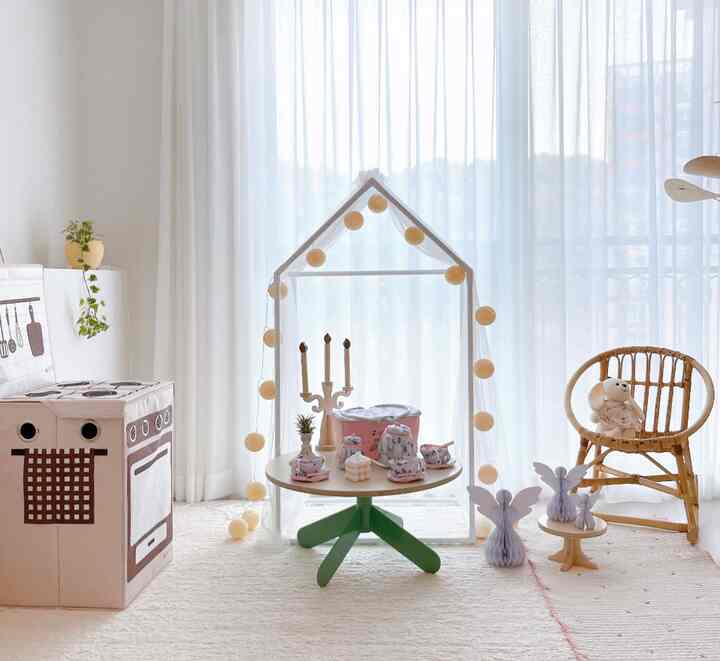 明るく澄んだ白とパステルトーンの子供部屋、メッシュキャノピーが中央の低いテーブルとおもちゃの上に配置され、ナチュラルな木製チェアが居心地の良い空間を演出