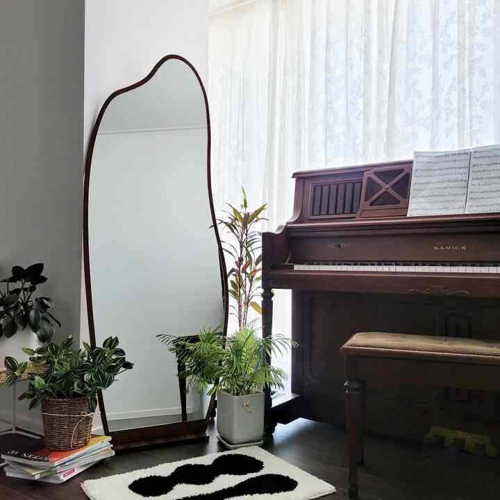 Natural wood tones and white curtains blend in a living room space featuring an irregular full-length mirror and walnut piano with cozy atmosphere