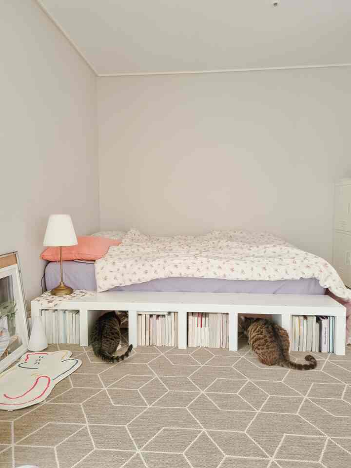 White and beige toned bedroom featuring a bed with a bookshelf underneath, a cat-shaped rug on floor, and two cats resting comfortably