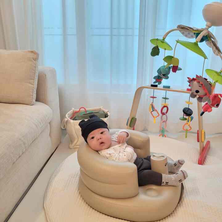Beige and white toned living room featuring a baby seated in a beige infant chair, with a mobile and baby play mat arranged in a cozy setting