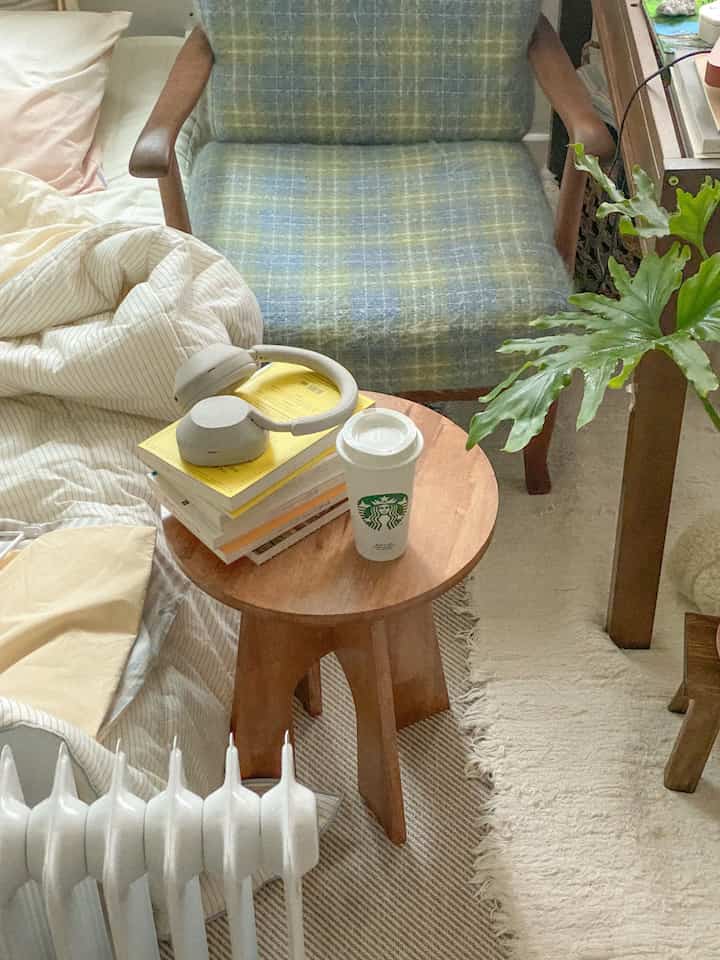 Natural tone bedroom space featuring a plaid sofa, wooden stool, desk, and plant creating a cozy vibe