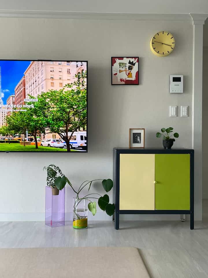 White and yellow toned living room featuring a TV, yellow-green cabinet, wall clock, and plants in a clean modern setting
