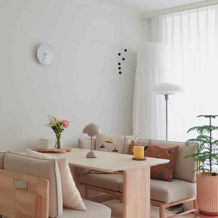 Bright beige and wood-tone living room featuring a corner sofa, large table, white curtains, and a simple table lamp in a cozy Nordic style