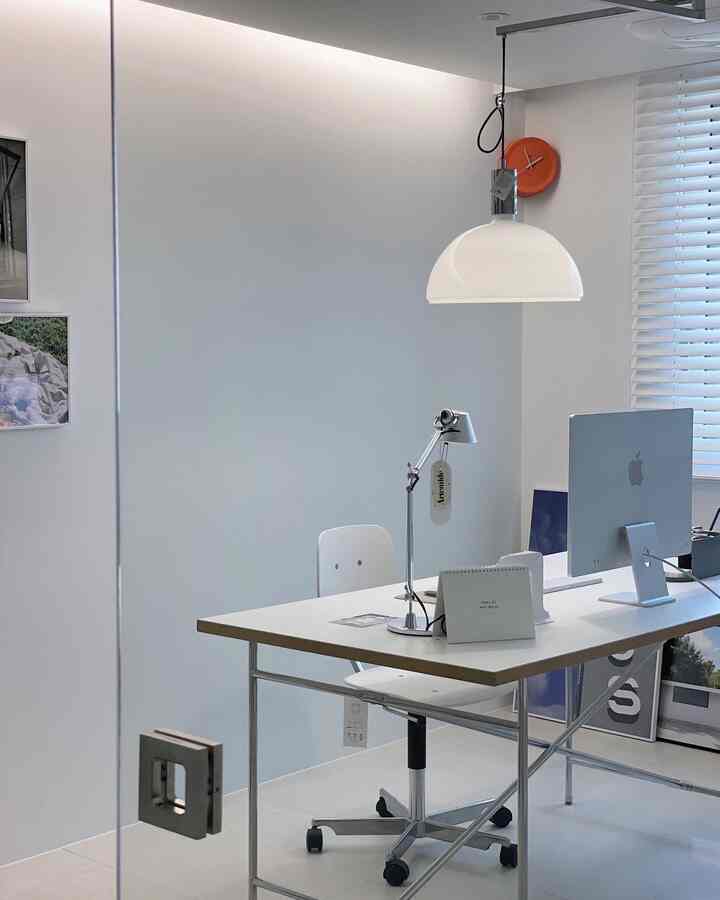 White-toned small home office featuring a simple desk, computer, table lamp, and pendant light in a clean interior