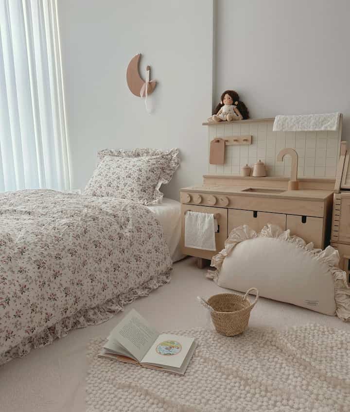 ベージュと白の色調の子供部屋、花柄の寝具と木製のおもちゃのキッチンが配置された暖かな印象の空間