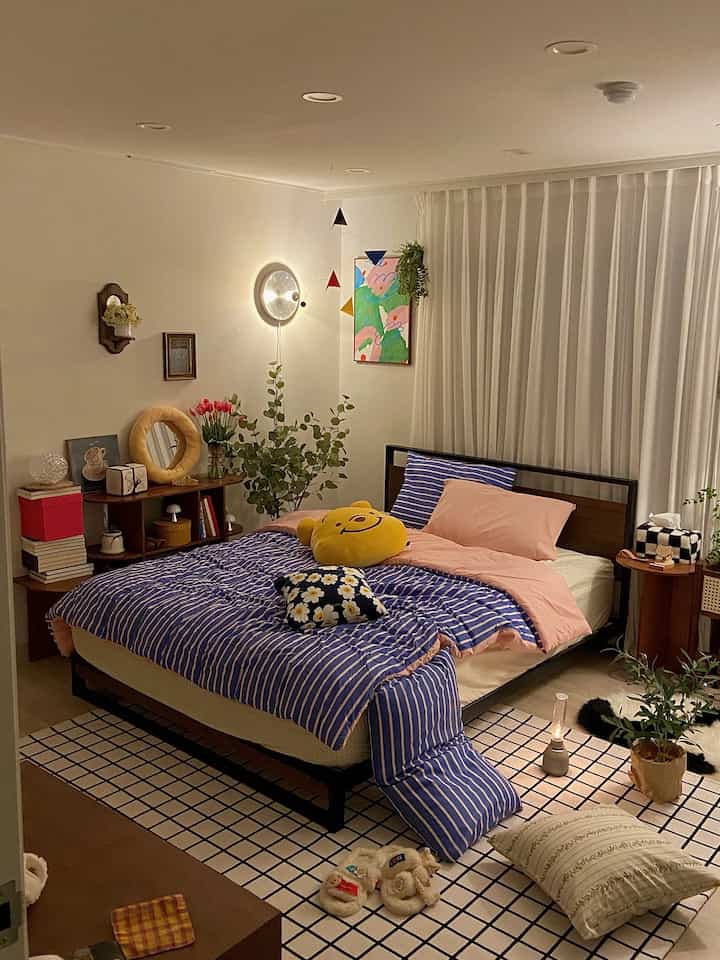 温かみのある色調の寝室、青のストライプ布団と木目調のベッドフレーム、白いカーテンと格子模様のラグが配置された雰囲気のあるインテリア