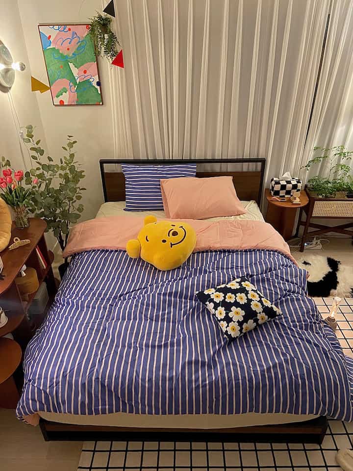 A bedroom with white curtains and blue striped bedding featuring natural wood furniture and indoor plants in a cozy setting