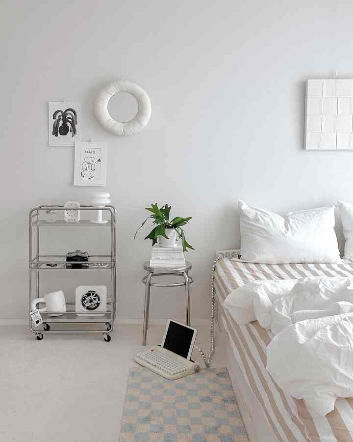 明るいホワイトトーンの寝室、ストライプの寝具とステンレススチールのトロリー、そしてグリーンプラントが配置されたミニマルな空間