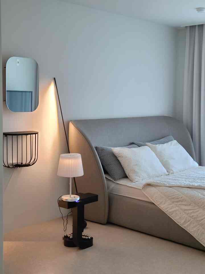 Monotone and white bedroom featuring a gray bed, nightstand, floor lamp, and wall mirror in a simple setting