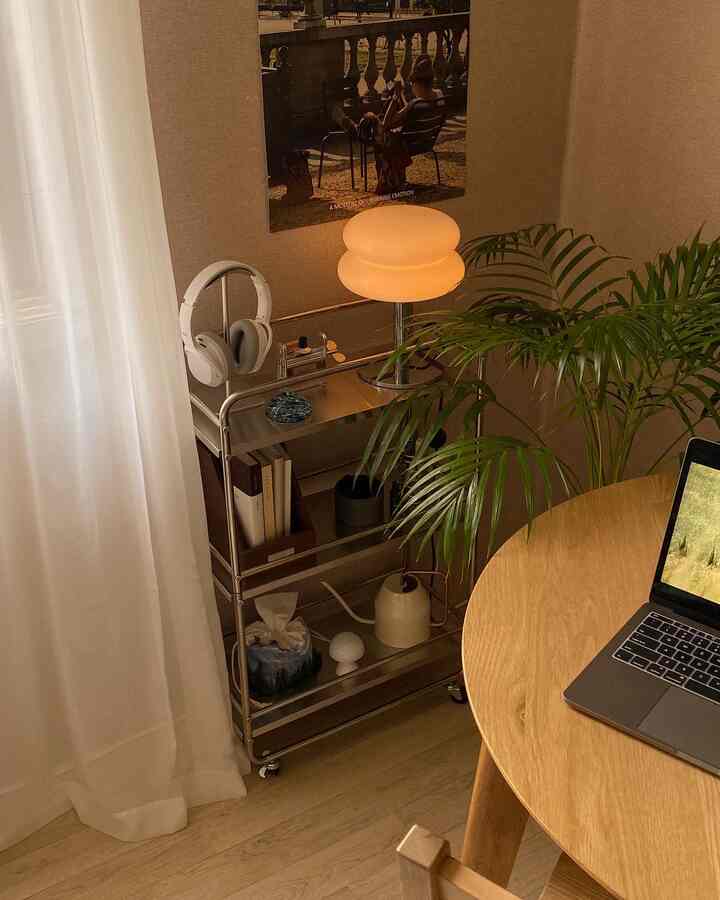 ベージュトーンの壁と木目調の丸テーブル、コーナーに置かれた金属製の収納カートと植物がある居心地の良いホームオフィスの空間