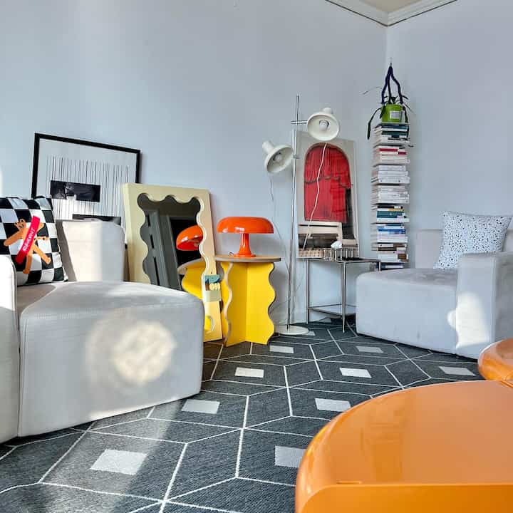 Living room in white and orange tones featuring two sofas, a yellow side table, floor lamp, and stacks of books, creating a clean and bright atmosphere