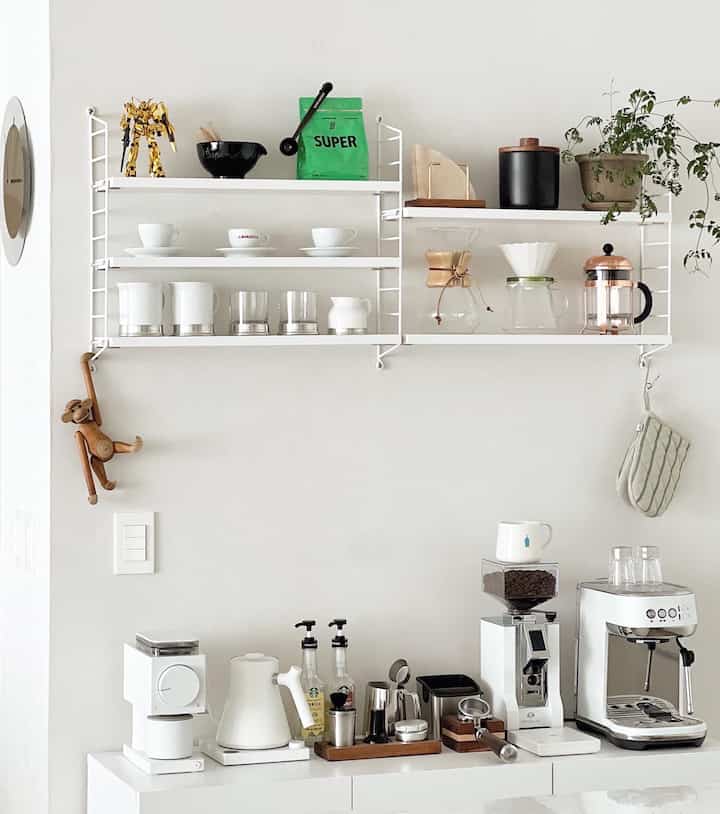 Bright white-toned kitchen featuring wall shelves and various coffee machines and cups, showcasing a clean modern home cafe