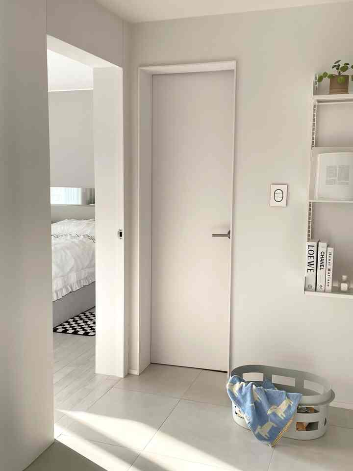 Bright white and light gray toned entrance area with adjacent bedroom, featuring wall shelving and laundry basket in a clean Nordic style space