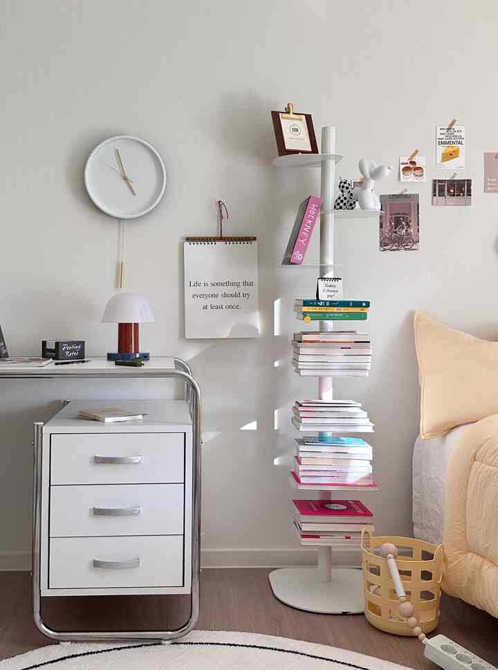 White-toned bedroom featuring a desk, silent wall clock, and bookshelf neatly arranged in a simple space