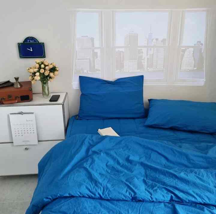 Cozy bedroom with blue bedding, white walls, retro clock, and vase on dresser