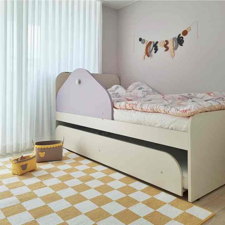 Modern kids' room with yellow checkerboard rug and white curtains, featuring lavender-accented bed and storage baskets