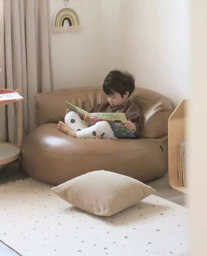 Natural-toned kids' room with brown bean bag and beige cushion, featuring a child comfortably reading a book